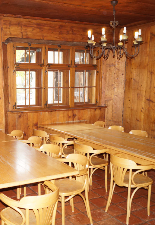 Großer Salon im Hostel Feldkirch mit langen Holztischen, vielen Sitzplätzen, Fenstern und rustikalen Kronleuchtern