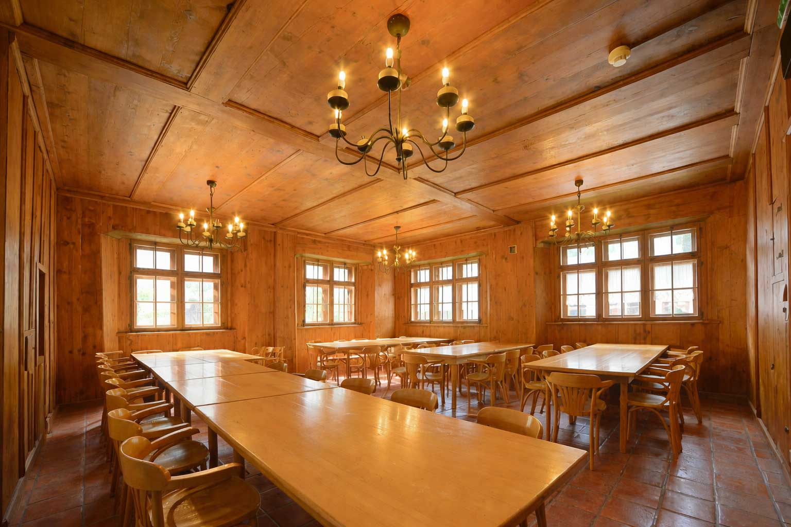Großer Salon im Hostel Feldkirch mit langen Holztischen, vielen Sitzplätzen, großen Fenstern und mehreren Kronleuchtern in warmem Holzambiente